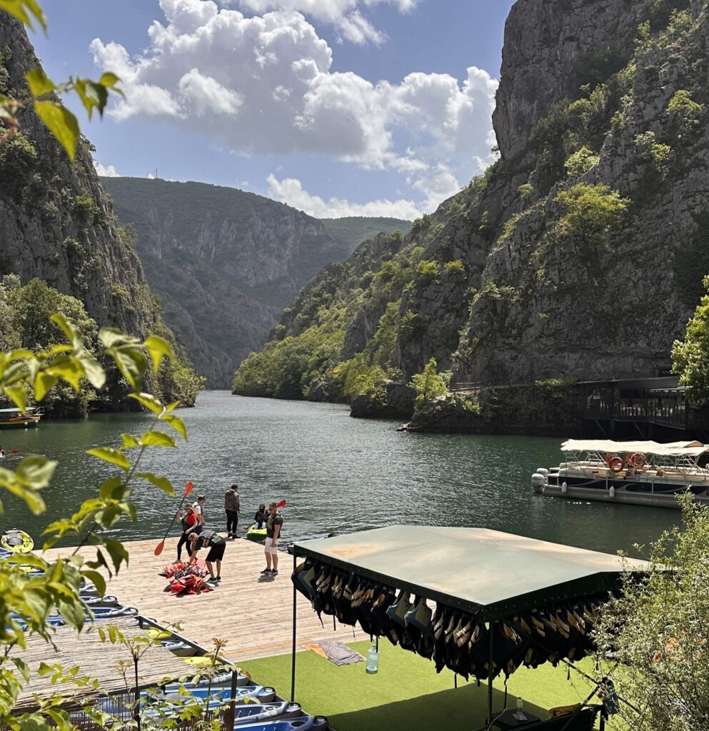 Blick auf den Matka Canyon mit blauem Wasser zwischen grünen Bergen und einem Steg mit Boot und Menschen im Vordergrund