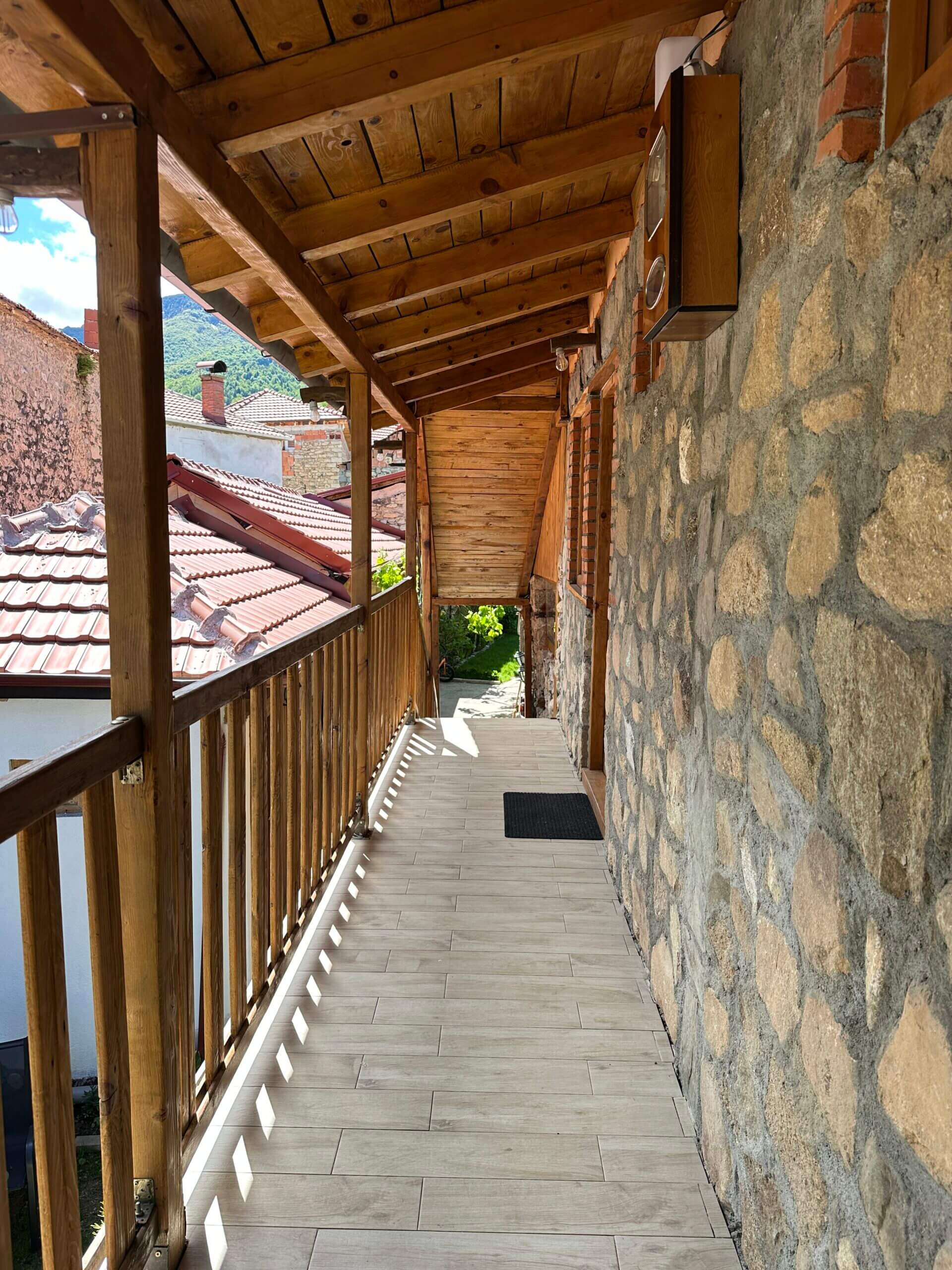 Überdachter Holzbalkon einer traditionellen Unterkunft mit Steinmauer und Blick auf Ziegeldächer im Ort