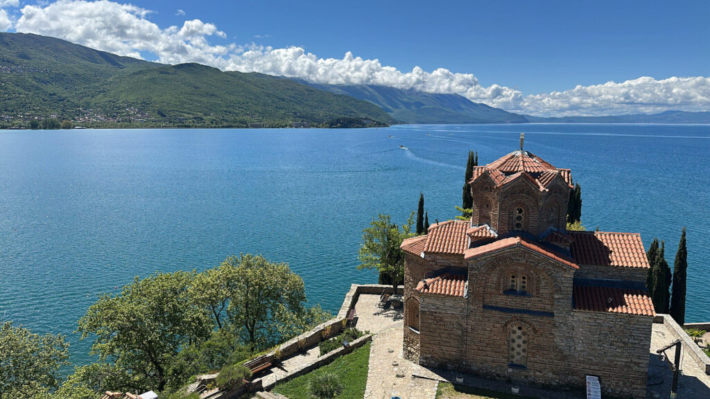 Blick auf den Ohrid-See mit der Kirche des Heiligen Johannes von Kaneo im Vordergrund, strahlend blauem Himmel und Wolken, die sich in den Bergen verhangen haben