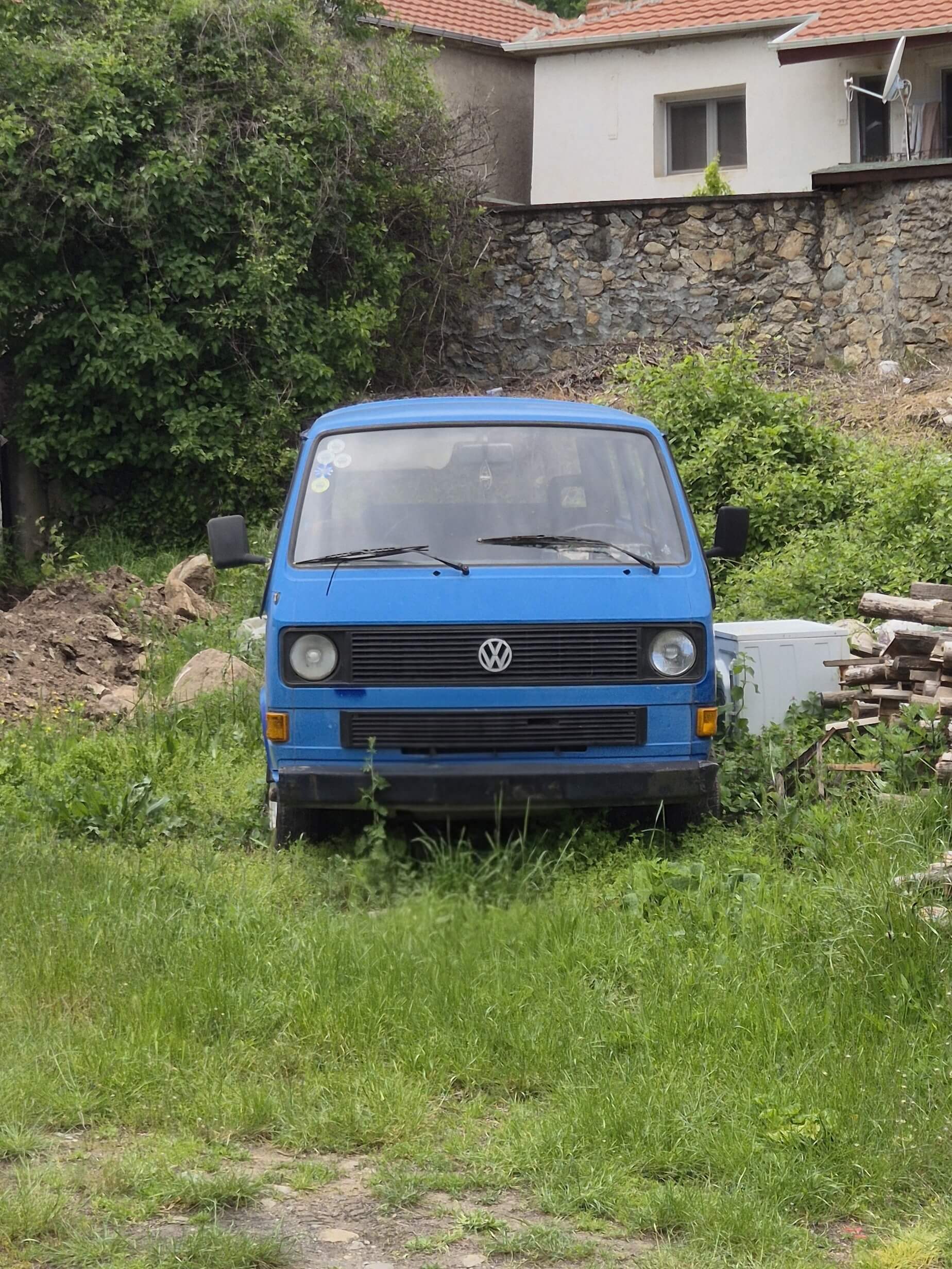 Blauer VW Bus auf grüner Wiese vor Steinmauer und Wohnhaus in Nordmazedonien