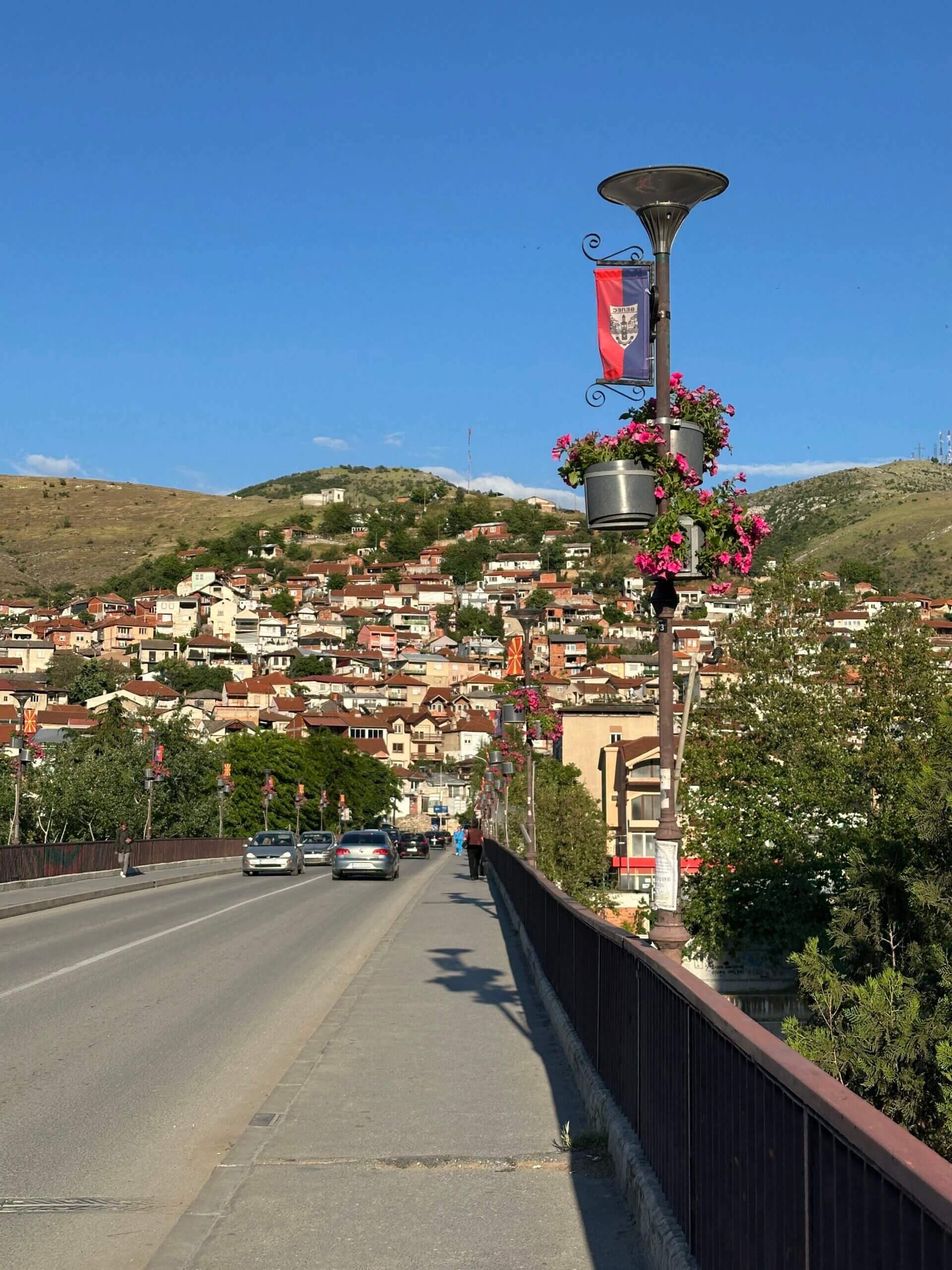Brücke mit blumenbehangenden Laternen, Autoverkehr und Blick auf Häuser am Hang in Veles